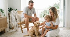 A happy young family—mother, father, and toddler daughter—sitting on a textured rug in a sunlit, coastal-style living room and petting their apricot Waterhill cavoodle puppy. The room features modern rattan furniture and a balcony view of green foliage and the ocean.