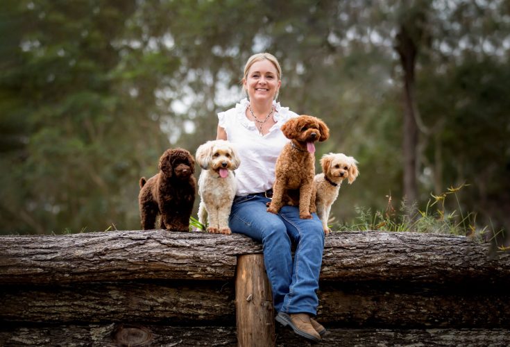 Waterhill Cavoodles breeder, Sarah Orchard, sitting on a rustic log at their 80-acre Sunshine Coast hinterland property, surrounded by four healthy and happy Cavoodle puppies in chocolate, cream, and apricot.